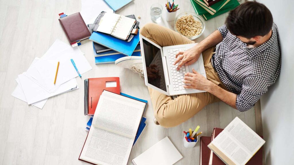 Young man sitting on the floor using laptop surrounded with great number of different things to search for the best shipping company